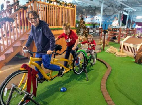 Three children ride a yellow tandem bike indoors, surrounded by a colorful play area with decorations and greenery.