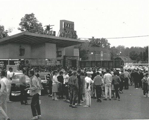 A large crowd lines up outside Paul’s Frozen Custard, with vintage cars parked nearby and trees in the background.