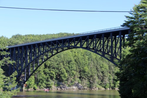 A large black arch bridge spans a river, surrounded by lush green trees and a clear blue sky.