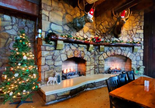Cozy holiday scene with two fireplaces, decorated stone wall, Christmas tree, and festive deer heads wearing Santa hats.