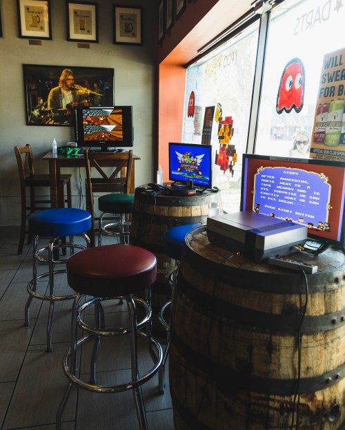 A cozy gaming corner with retro consoles on barrels, two TVs displaying classic games, and colorful stools.