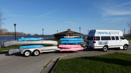A trailer loaded with colorful kayaks parked near a lake, with a rental van nearby and a gazebo in the background.