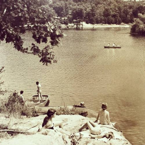 A black-and-white photo of people by a lake, some in boats, others relaxing on rocks by the water. Trees in the background.