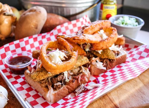 Two sandwiches piled with pulled pork, coleslaw, fried pickles, and onion rings, served with dipping sauce on a checkered tray.