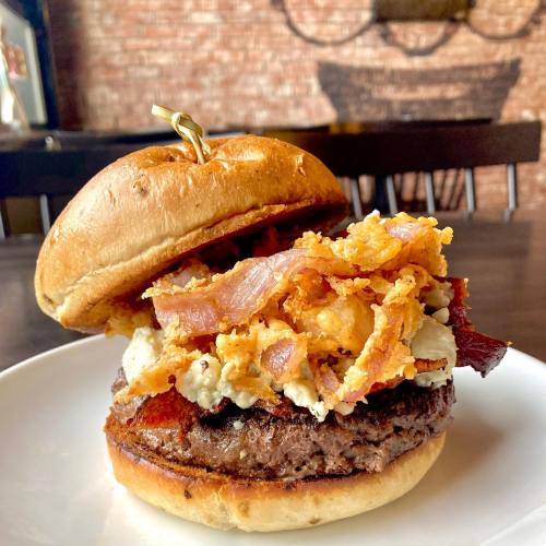 A close-up of a burger with a beef patty, crispy bacon, and onion rings on a toasted bun, served on a white plate.
