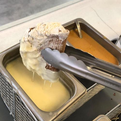 A cone of ice cream being dipped into a container of white chocolate, with some chocolate dripping off.