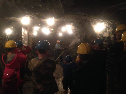A group of people in hard hats watches a miner working in a dimly lit underground mine.