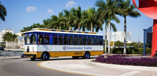 A colorful trolley bus labeled "Sweetwater Trolley" turns at a roundabout surrounded by palm trees and buildings.