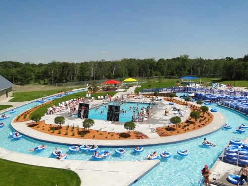 A sunny water park scene with a lazy river, pool area, and people enjoying various activities. Colorful umbrellas dot the landscape.