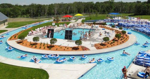 A vibrant water park scene with a circular pool, lazy river, and colorful umbrellas, filled with people enjoying the sun.