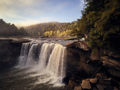 A stunning waterfall cascades over rocky cliffs, surrounded by lush greenery and misty morning light.