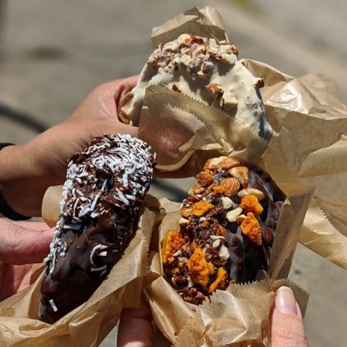 Three hands hold ice cream bars coated in chocolate and toppings, wrapped in parchment paper.