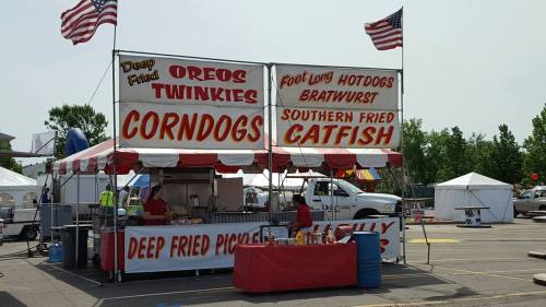 A food stand at a fair featuring deep-fried treats like Oreos, Twinkies, corndogs, and catfish, with American flags.
