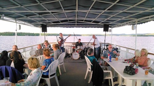 A band performs on a boat deck while guests enjoy drinks and conversation by the water.