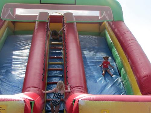 Children playing on a colorful inflatable slide, with one climbing up and two sliding down. Bright sunny day.