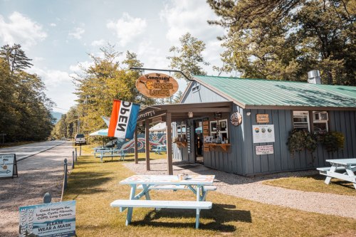A small, rustic building with a green roof and outdoor seating, surrounded by trees and a road, with an "Open" sign.