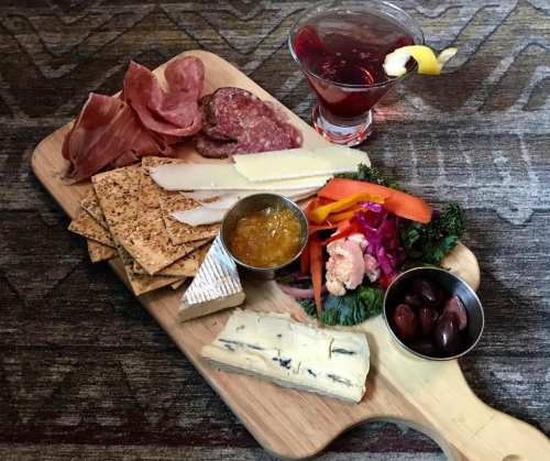 A wooden platter with assorted meats, cheeses, crackers, olives, and a cocktail garnished with a lemon slice.