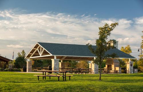 A spacious picnic shelter with stone pillars, surrounded by green grass and trees under a blue sky with clouds.