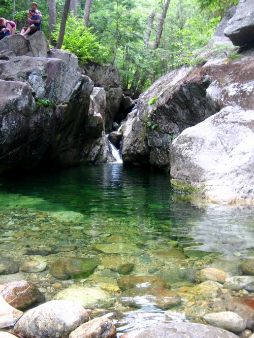 A serene natural pool surrounded by large rocks and lush greenery, with a small waterfall in the background.