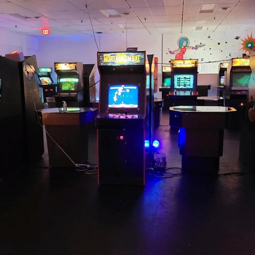 A dimly lit arcade featuring several vintage game machines, with a "Mortal Kombat" cabinet in the foreground.