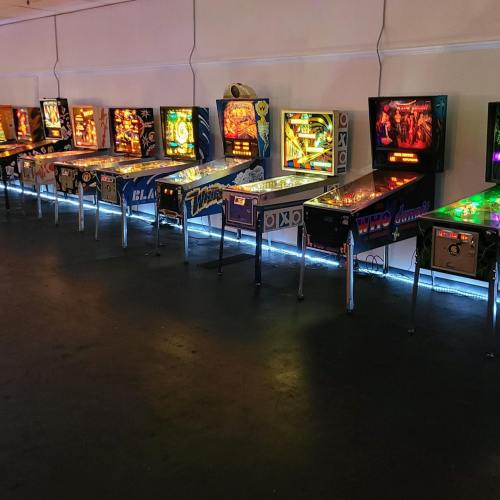 A row of colorful pinball machines illuminated in a dimly lit arcade setting.