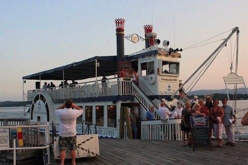 A riverboat docked at sunset, with people on the deck and pier, enjoying the scenic view.
