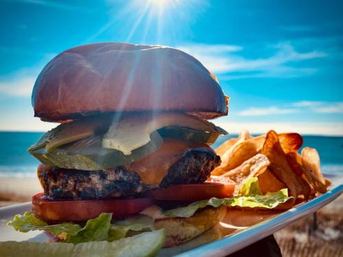 A delicious burger with lettuce, tomato, pickles, and cheese, served with chips, against a sunny beach backdrop.
