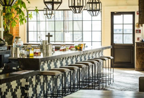A stylish bar with a patterned counter, stools, and decorative lighting, featuring fresh fruits and a large window.