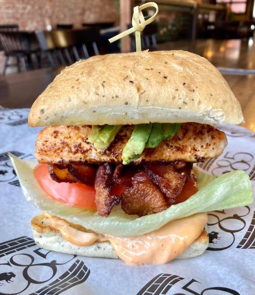 A delicious sandwich with grilled chicken, bacon, lettuce, tomato, and avocado on a whole grain bun, served on parchment paper.
