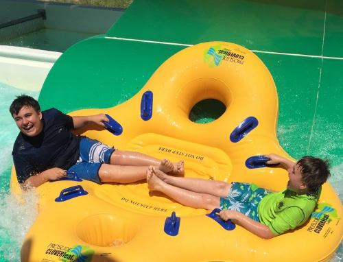 Two boys laughing on a large yellow inflatable raft at a water park, splashing water around them.
