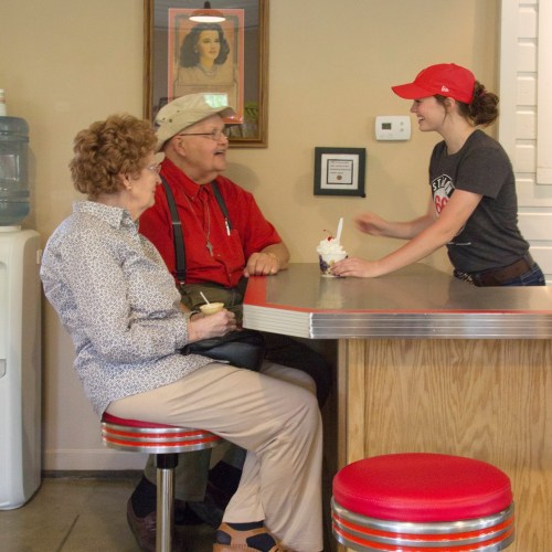 A woman in a red cap serves ice cream to an elderly couple sitting at a diner counter.