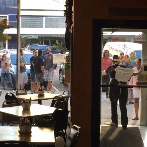 A view from inside a restaurant showing people waiting outside and a staff member at the entrance.
