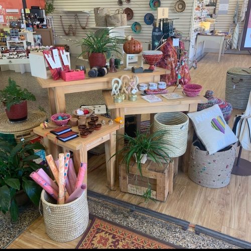 A wooden display table with various decorative items, plants, and woven baskets in a vibrant shop setting.