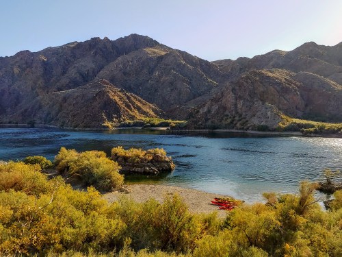 A serene river scene with mountains in the background, featuring a sandy shore and kayaks on the water.