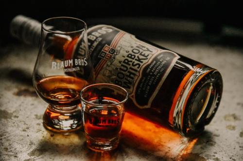 A bottle of bourbon whiskey with two glasses, one filled with whiskey, on a textured surface.