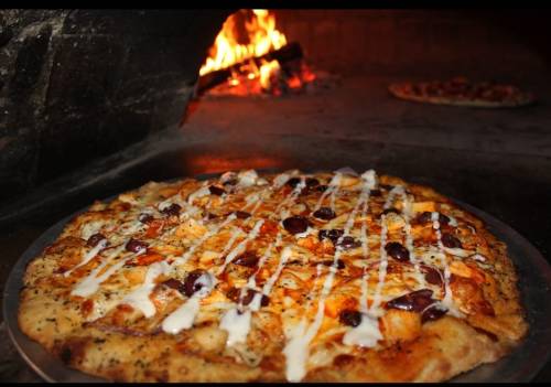A freshly baked pizza with toppings and drizzled sauce, placed in a wood-fired oven with flames in the background.
