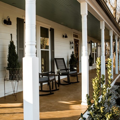 A cozy porch with rocking chairs, potted plants, and white columns, featuring a blue ceiling and warm sunlight.