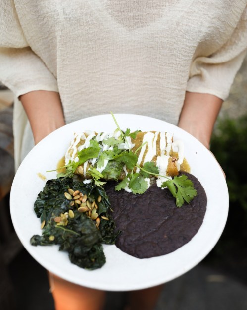 A person holds a plate with enchiladas, black beans, and sautéed greens, garnished with cilantro and drizzled sauce.