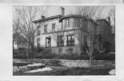 Historic two-story house with a curved balcony, surrounded by trees and a stone wall, in black and white.