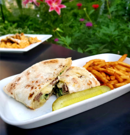 A grilled sandwich with greens and cheese, served with fries and a pickle, against a backdrop of colorful flowers.