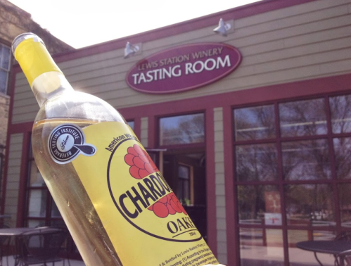 A bottle of Chardonnay in front of the Lewis Station Winery Tasting Room, showcasing the winery's sign.
