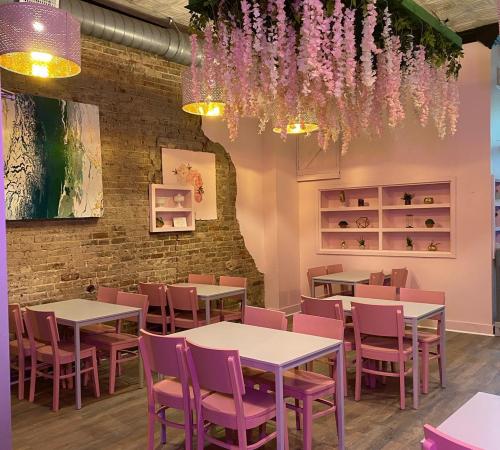 A cozy café with pink chairs and tables, adorned with hanging flowers and a colorful wall art piece.