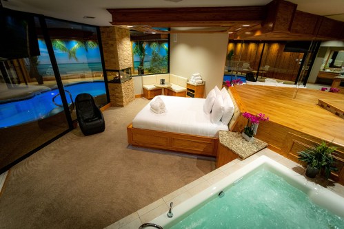 Luxurious hotel room with a king bed, hot tub, and views of a pool and palm trees through large windows.