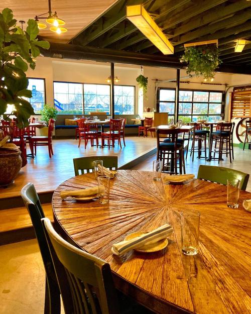 A wooden table with empty glasses and utensils, surrounded by a spacious restaurant with green plants and seating areas.