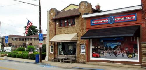 A brick building featuring a mural, with an American flag and benches outside, in a small town setting.