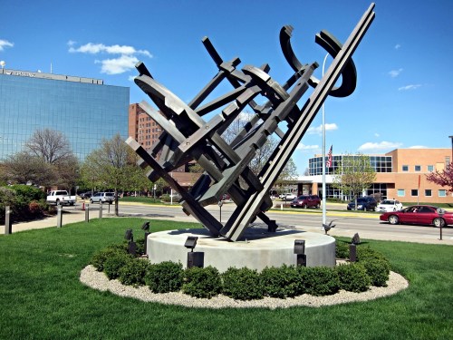 Abstract metal sculpture on a circular base, surrounded by grass and trees, with buildings in the background under a blue sky.