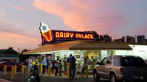 A colorful sunset over Dairy Palace, with people gathered outside enjoying ice cream. Neon sign glowing brightly.