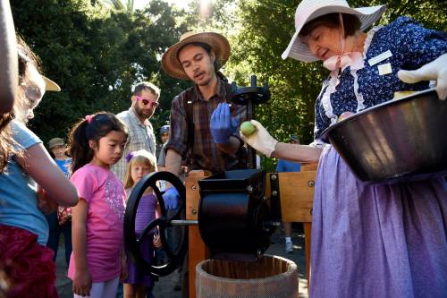A woman in historical attire demonstrates a fruit press to children and adults at an outdoor event.