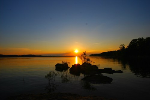 Sunset over a calm lake, with silhouettes of rocks and trees against a vibrant orange and blue sky.
