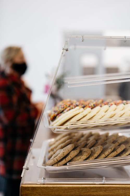 A display case with various cookies, including colorful frosted ones, in a bakery setting. A person is blurred in the background.
